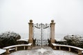 A closed black metal gate with concrete pillars surrounded by green plants covered in snow Royalty Free Stock Photo