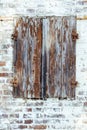 Closed brown red peeling wooden window with rusted metal hinges on a grungy white brick wall of an old abandoned derelict barn