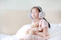 Closed eyes little Asian child girl wearing winter earmuffs and embracing teddy bear while sitting on the bed at home Royalty Free Stock Photo