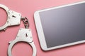 Closed handcuffs and tablet on a pink background. Computer crime concept