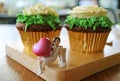 Closed up mini angel holding pink big heart figurine with a pair of flower shaped whipped cream chocolate cupcakes in background