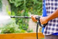 Closeup arms of man wearing square pattern blue and white shirt holding high pressure water gun, pointing towards green garden