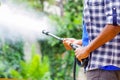 Closeup arms of man wearing square pattern blue and white shirt holding high pressure water gun, pointing towards green garden