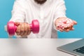 Closeup bearded man holding out hands with pink dumbbell and sweet donut with icing to camera sitting at workplace with laptop,