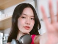 Closeup of beautiful Asian woman wearing headphones around her neck and making selfie shot on mobile phone stands in kitchen.