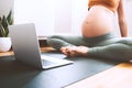 Closeup belly of pregnant woman practicing yoga online with laptop