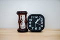 Closeup black alarm clock for decorate next to hourglass, wooden desk front concrete wall background. Royalty Free Stock Photo