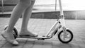 Closeup black and white photo of female legs standing and riding on kick scooter on the road