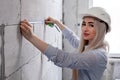 Closeup blonde girl designer foreman in white construction helmet measures the grey wall of sibit by tape measure in house under