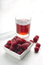 Vertical image.Glass of raspberry drink, square bowl of raspberries o the white marble table