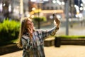 Closeup cheerful girl taking selfie photo by mobile phone on night city street. Portrait of happy woman showing victory sign to Royalty Free Stock Photo