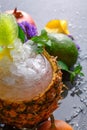 Closeup of coctail in a glass bowl in a pineapple, flowers, leaves of mint, pear, pomegranate on a bluerred background.