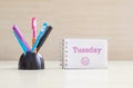 Closeup color pen with black ceramic desk tidy for pen and pink tuesday word in white page and frown face emotion at the note book