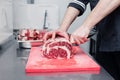 Closeup cook`s hand cuts ribeye marbled beef steak with sharp knife on red plastic cutting board on metal table in restaurant