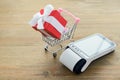 Closeup of credit card terminal with touchscreen display and red gift box with white bow in miniature shopping cart on a