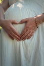 Closeup cute composition with a pregnant young woman wearing white dress makes a heart shaped symbol with her hands on her belly. Royalty Free Stock Photo