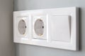 A closeup diagonal view of a group of white european electrical outlets and a switch located on a painted gray wall