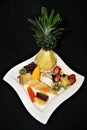Closeup display of assorted sliced fruits and cheeses with sculpted Pineapple on a white plate with a black background