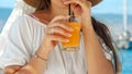 Closeup of elegant woman holding glass of orange juice and drinking it through straw while relaxing on wooden sea pier Royalty Free Stock Photo