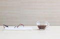 Closeup eyeglasses on white book with black coffee in transparent cup of coffee on blurred wooden desk and wall textured