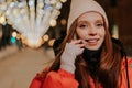 Closeup face of pretty young woman in hat and winter jacket talking holding smartphone standing on snow city street on Royalty Free Stock Photo