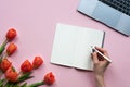 Closeup of female hand writing on a notebook. Pink background with laptop and tulips. Copy space. Royalty Free Stock Photo