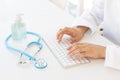 Closeup of female hands typing on computer keyboard, gel hand sanitizer and stethoscope. Online medical consultation and virus pro