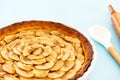 Closeup on a French apple tart aside a cooking spoon and a rolling pin on a light blue background. Side view