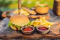 Closeup of fresh burger with French fries on wooden table with bowls of tomato sauce. lifestyle food Royalty Free Stock Photo