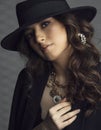 Closeup front portrait of young brunette woman in black jacket and hat with makeup, curly hair, over grey background. Royalty Free Stock Photo