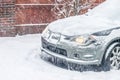 Closeup of front of silver car in snowstorm with lights on but covered in snow and icicles