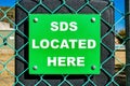 Closeup of green square SDS LOCATED HERE sign on turquoise mesh wire fence against black surface Royalty Free Stock Photo