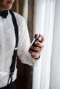 Closeup of the groom in shirt and suspenders stands by the window holding a bottle of male perfume in her hand