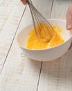 Closeup of a hand beating an egg on a bowl with an egg beater on a white plank table