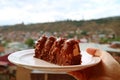 Closeup hand holding a plate of chocolate with caramel mousse cake against blurry city aerial view Royalty Free Stock Photo