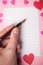 Hand of man writing with vintage pen on spirales note book page with paper hearts on pink background Royalty Free Stock Photo