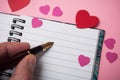 hand of man writing with vintage pen on spirales note book with paper hearts on pink background Royalty Free Stock Photo
