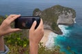 Closeup hands with mobile phone doing photo of Kelingking Beach cliffs in Nusa Penida, Bali