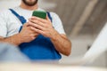 Closeup of hands of repairman in workwear texting, using smartphone, standing indoors during renovation work