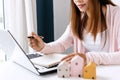 Closeup of happy young Asian woman searching home information online in a laptop and writing on notebook on a table at home.