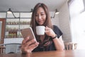 Closeup image of a beautiful Asian woman with smiley face holding and using smart phone while drinking hot coffee on wooden table Royalty Free Stock Photo