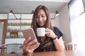 A beautiful Asian woman with smiley face holding and using smart phone while drinking hot coffee on wooden table in modern cafe Royalty Free Stock Photo