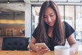 A beautiful Asian woman with smiley face holding and using smart phone with white coffee cup on wooden table in modern loft cafe Royalty Free Stock Photo