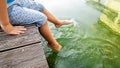 Closeup image of child sitting on the wooden pier at tiver and holding feet in water. Kids playing and splashing water Royalty Free Stock Photo