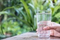 Closeup image of a hand holding a glass of cold water on wooden table with green nature