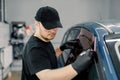 Closeup image of a handsome car mechanic worker, wearing black uniform, attaching tinting foil to car window in