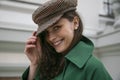 Closeup image of joyful elegant young woman in hat and green coat smiling looking at camera, building background