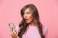 Closeup image of puzzled woman having beautiful long hair looking at smartphone in hand with bulging eyes, isolated over pink