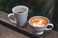Closeup image of two cups of black coffee and latte with heart latte art on wooden bench in green nature Royalty Free Stock Photo