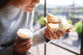 A woman holding and eating whole wheat sandwich and coffee in the morning